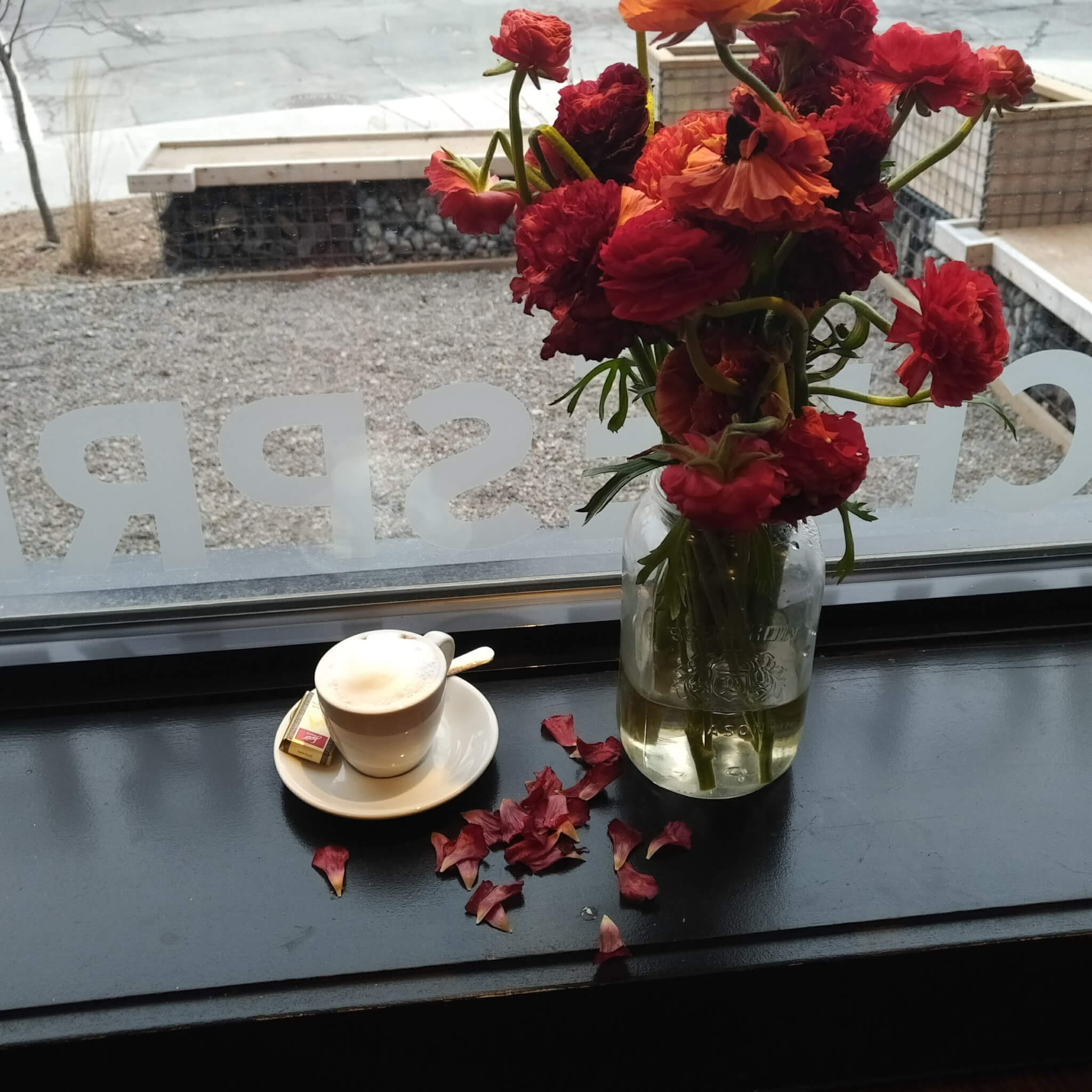 a single macchiato in a white ceramic espresso cup and saucer. A bouquet of red flowers is beside and spilling petals onto the windowsill.